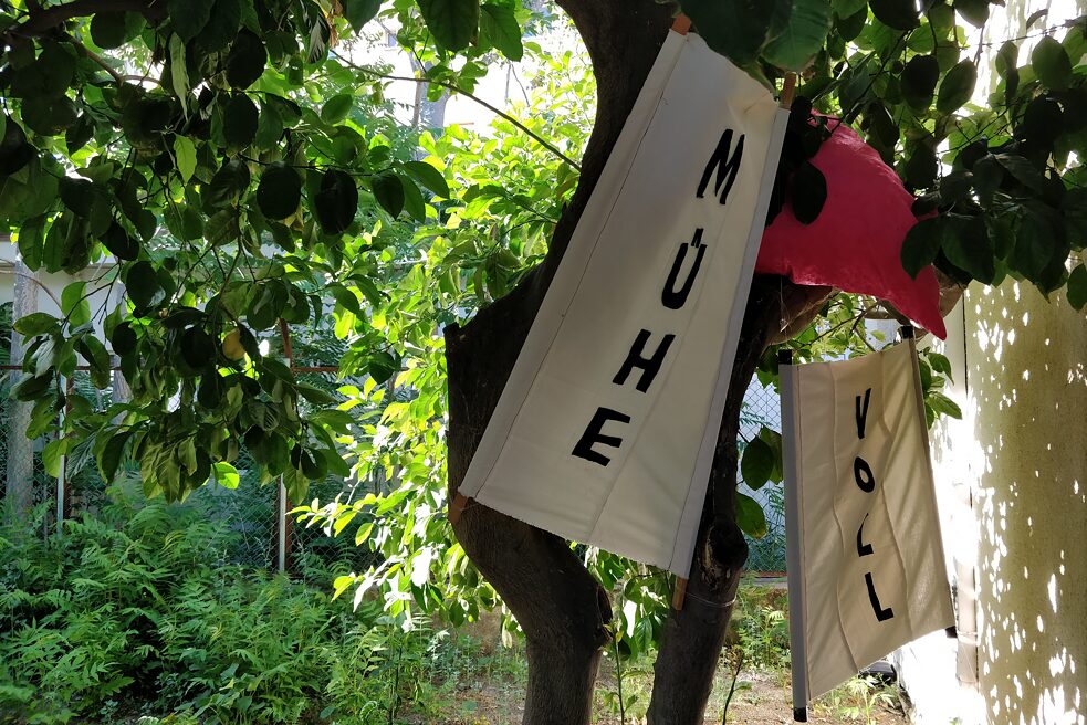 The image shows a tree in which two white fabric banners are attached. One banner says Mühe and the banner further back in the picture says Voll. Between them is a pink colored installation.