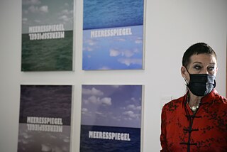 On the right side of the picture you can see a woman (Dagmar Glausnitzer-Smith). Next to her hang four pictures, in two rows, showing the sea and the horizon. On all the pictures is the word Meeresspiegel.