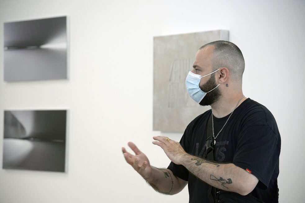 In the foreground you can see the side profile of a man (Stelios Kallinikou). He is wearing a black T-shirt and has tattoos. He gestures with his hands. In the background you can see three works of art.