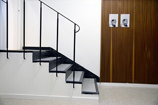 The right half of the picture is occupied by a staircase. Next to it, two portraits of monkeys hang on a wooden paneling on the wall.