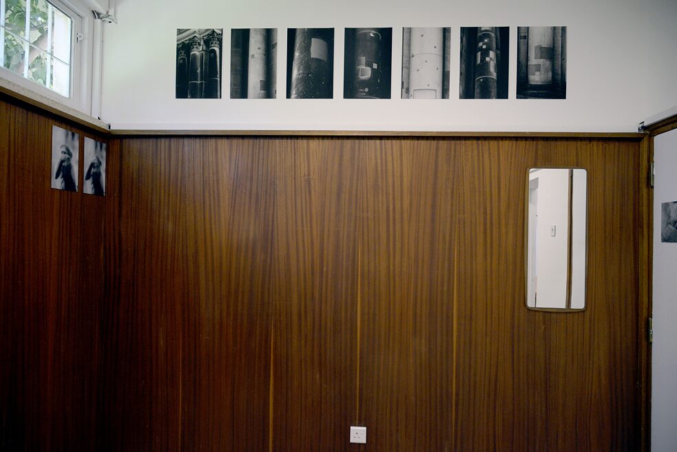 In the picture you can see a room, which is two-thirds covered with wood. On the wall hangs a mirror in the right corner. Above the wooden paneling hangs a series of pictures. On the left wall, in the right upper corner hang two more pictures.