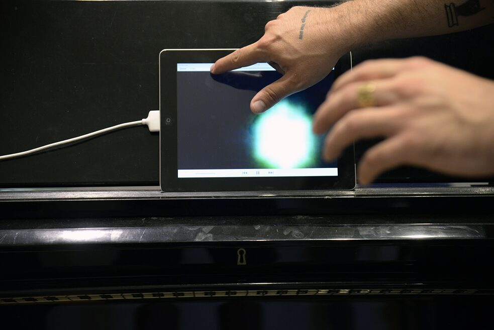 Das Bild zeigt ein Tablett, das auf dem Notenpapierhalter eines Klaviers steht. Das Tablett wird von einer Hand bedient. Davor sieht man noch eine weitere leicht verschwommene Hand. Auf dem Tablett wird eine Datei abgespielt.