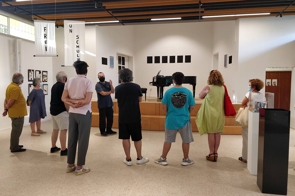 Μια ομάδα 8 ατόμων στέκονται σε ημικύκλιο γύρω από έναν άνδρα (Στέλιος Καλλίνκου) ο οποίος μιλά. Γύρω από τους ανθρώπους, φαίνονται διάφορα έργα τέχνης.