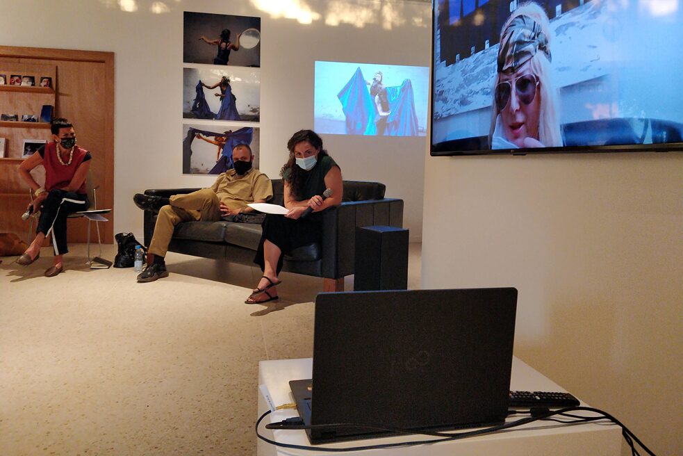 Im Vordergrund des Bildes ist ein laptop zu shene. Daneben hängt ein Bildschirm auf dem eine Künstlerin zu sehen ist. Im Hintergrund an den Wänden sind weitere Kunstwerke. Vor diesen steht ein Sofa auf dem zwei Personen sitzen und ein Stuhl mit einer weiteren Person.