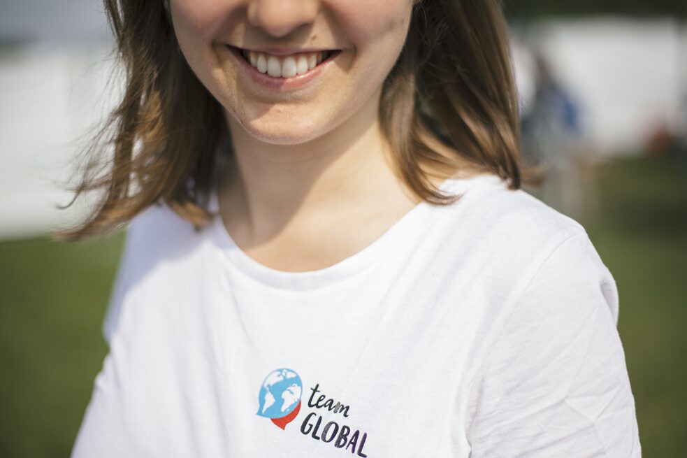 Nahaufnahme des Gesichts einer Mitarbeiter*in der Organisation "Team Global". Auf dem T-Shirt steht neben einer Grafik der Erdkugel "Team Global".