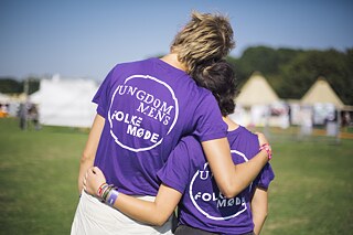 Im Vordergrund stehen zwei Helfer*innen von Ungdommens Folkemøde mit dem Rücken zu Kamera. Sie tragen T-Shirts mit dem Text "Ungdommens Folkemøde" und halten die Arme umeinander. 