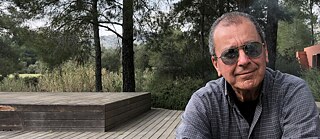 A middle-aged man with grey hair, sunglasses and a plaid shirt is seated in the lower right of the photo. Behind him can be seen a wooden floor with a raised platform and a row of pine trees.