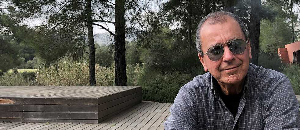 Ein Mann mittleren Alters mit grauem Haar, Sonnenbrille und kariertem Hemd sitzt unten rechts auf dem Foto. Hinter ihm ist ein Holzboden mit einer erhöhten Plattform und einer Reihe von Kiefern zu sehen.