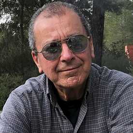Έναςμεσήλικας άντρας με γκρίζα μαλλιά, γυαλία ήλιου και καρώ πουκάμισο είναι καθιστός στο κάτω δεξί μέρος της φωτογραφίας. Πίσω του φαίνονται ένα ξύλινο δάπεδο με μια υπερυψωμένη εξέδρα και μια σειρά από πεύκα.