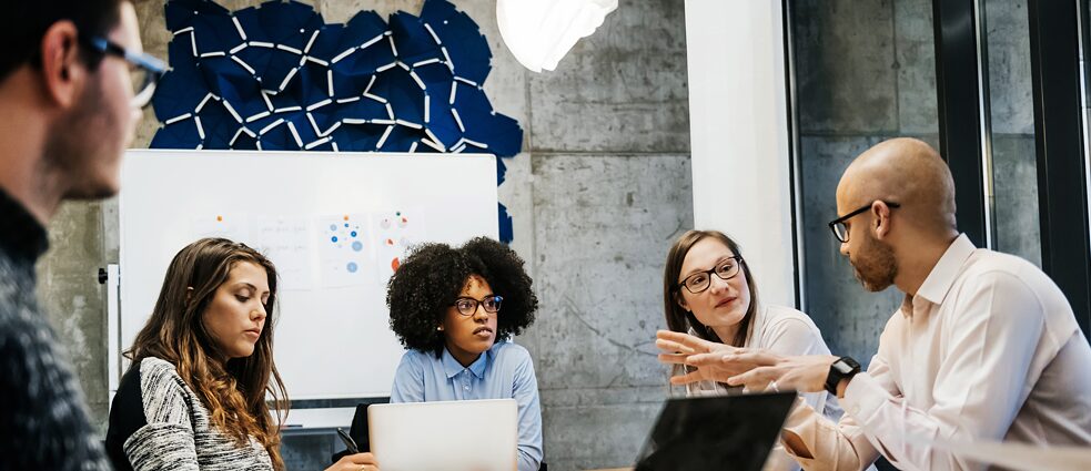 Junge Frauen und Männer sitzen zu einer Besprechung an einem Tisch. Ein Mann spricht und gestikuliert.