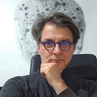 Portrait of a man (Achileas Kentonis). He is wearing a dark grey turtleneck jumper and round glasses. He is sitting on a desk chair. Behind him is a white wall with an artwork. With his right hand he grasps his chin.