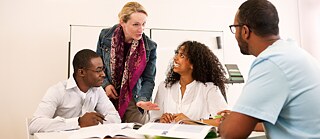 Zwei Kursteilnehmer und eine Kursteilnehmerin sitzen im Klassenraum, die Lehrerin steht bei ihnen am Tisch.