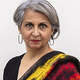 The picture shows a portrait of a woman. She is standing sideways and her head is turned towards the camera. She is wearing a black top. The shoulder facing the camera is covered with a colourful scarf. She wears gold creoles and red lipstick.