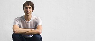 On the left of the picture a young man is sitting in a squat in front of a white wall, with his arms intertwined and elbows resting on his knees. He is wearing jeans and a white t-shirt.
