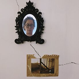 The painting shows a wall with a crack running through it. At the upper part of the crack a richly decorated frame hangs above the crack. In this frame a picture of a woman can be seen.  At the lower part of the crack hangs a yellowed paper with a photo of an old staircase on it.