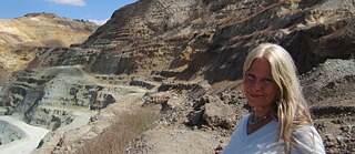 Two hills from the Skouriotissa mine can be seen in the background. A woman in a white blouse and long blonde hair is standing smiling in front of them on the right side of the picture.