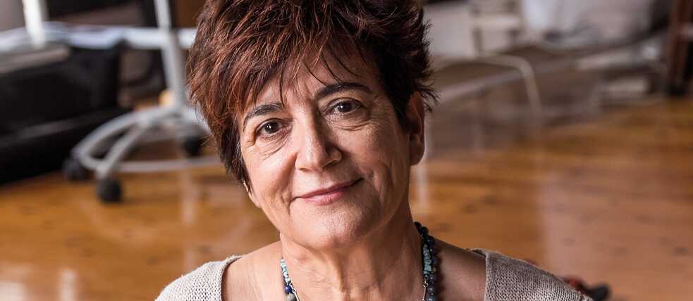 Μια γυναίκα (Αριάννα Οικονόμου) κάθεται σε ένα μπλε μαξιλάρι και στηρίζεται με το αριστερό της χέρι. Έχει κοντά κόκκινα βαμμένα μαλλιά, φοράει ένα κολιέ και ένα ανοιχτό γκρι πλεκτό πουλόβερ.