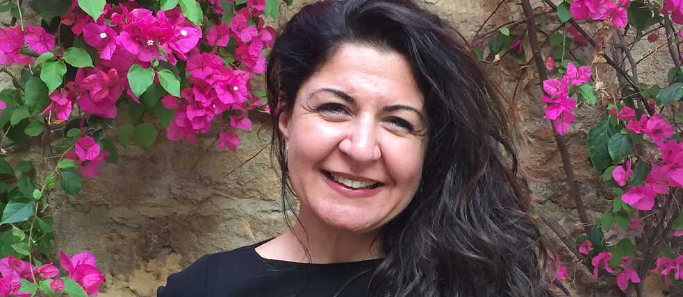 Siyah bluz giymiş bir kadın, begonvillerle dolu bir duvarın önünde duruyor ve gülümsüyor. Başı hafif sola eğik ve uzun siyah saçları sol omzuna dökülmüş.