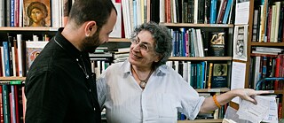 A young man with a black shirt, short hair and beard, and a middle-aged woman with short gray hair and a white shirt stand in front of bookshelves and among books. They look at each other and smile.