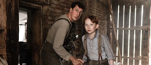 "Cam Oda" filminden bir sahne – Resimde bir ahırda bulunan iki kişi; solda bir yetişkin ve sağda bir erkek çocuk görülüyor. Her ikisi de dikkatle kamera yönüne bakıyor, birbirlerine doğru eğilmişler ve el ele tutuşmuşlar.