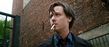 Σκηνή από την ταινία «Fabian – Going to the dogs» – Ένας νεαρός άντρας στέκεται με ένα τσιγάρο στο στόμα μπροστά από ένα κτίριο με τούβλα. Το βλέμμα του είναι στραμμένο στο πλάι. Φορά σακάκι, μπλε καρό πουκάμισο και κόκκινη γραβάτα.