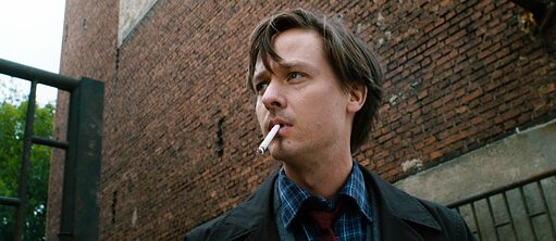 Scene from the film "Fabian - Going to the dogs" – A young man is standing with a cigarette in his mouth in front of a red-brick building. He is gazing into the distance. He is wearing a jacket, a blue checkered shirt and a red tie.