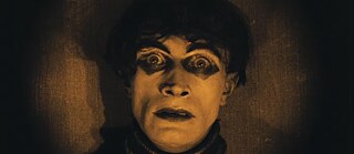 Das Bild zeigt einen jungen Mann in Großaufnahme. Er trägt einen Rollkragenpulli, sein Gesicht ist weiß geschminkt, unter seinen Augen sind zwei dunkle Dreiecke gemalt. Er schaut schreckensvoll in die Kamera.