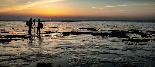 Scene from the film "Silence of the tides" – The picture shows a sunset at Wadden Sea. On the left the silhouettes of two people standing next to a camera mounted on a tripod can be seen. They are looking towards the sunset in the background of the picture.