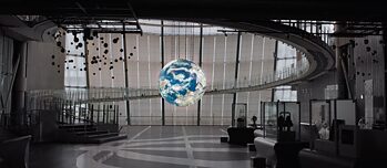 Scene from the film "Who we were" – A large room in grey tones. In the far back a glass wall with long windowblinds can be seen. In the middle of the room a very large colour 3D and illuminated globe is hanging from the ceiling. A staircase in the background leads to the next floor.
