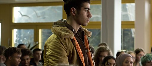 Scene from the film "Wet Dog" – A young man is standing in the middle of what seems like a school assembly, with his fellow students seated around him and some looking at him. He is wearing his long hear in a bun and a beige jacket with a hoody.