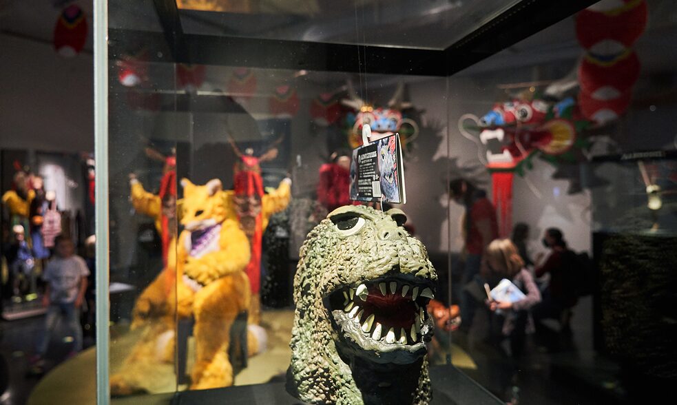 Close-up of a showcase with a dinosaur-like model of a head with more animal-like masks and figures in the background.