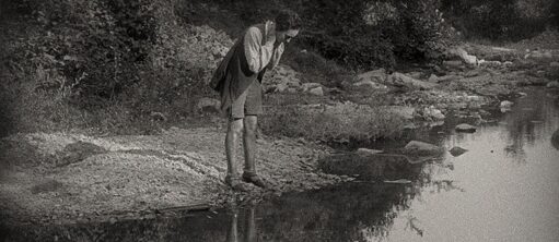 Une jeune femme debout au bord d'une rivière, penchée sur l'eau pour regarder son reflet.