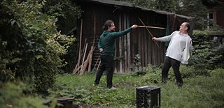 A young man and an older man are play fighting with sticks in a forest