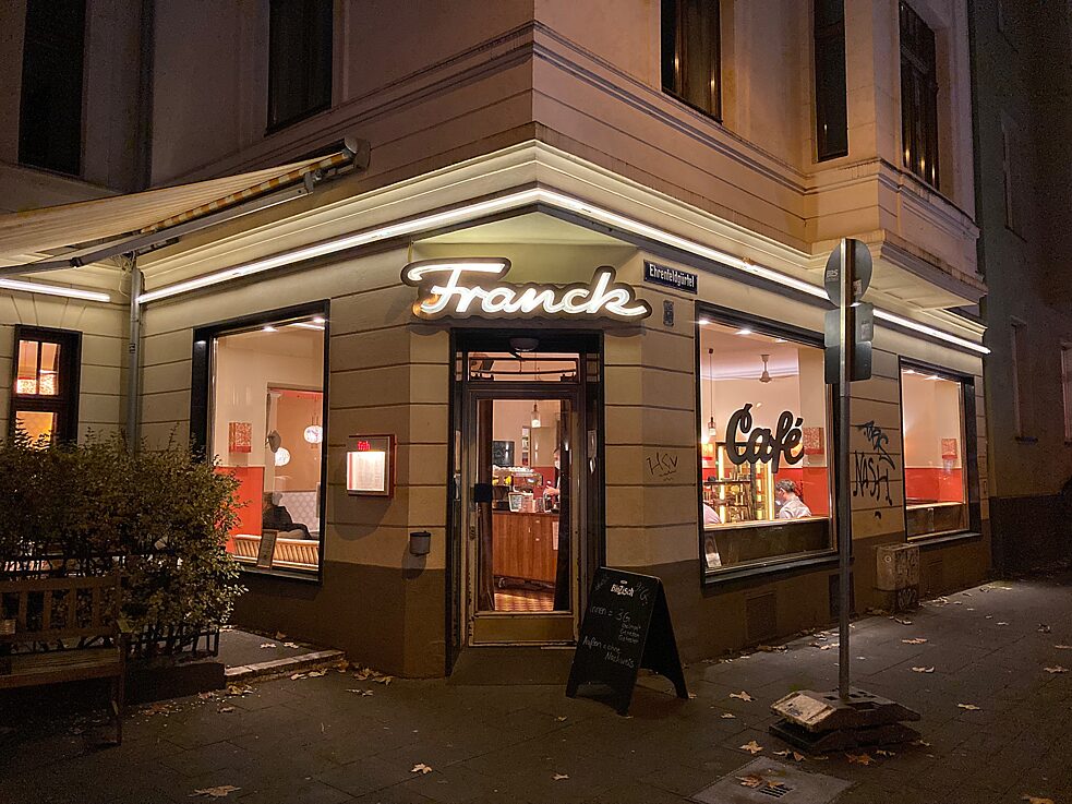 Café von außen: Straßenecke, über dem Eingang ein großes Leuchtschild in Retroschrift, auf dem "Franck" steht