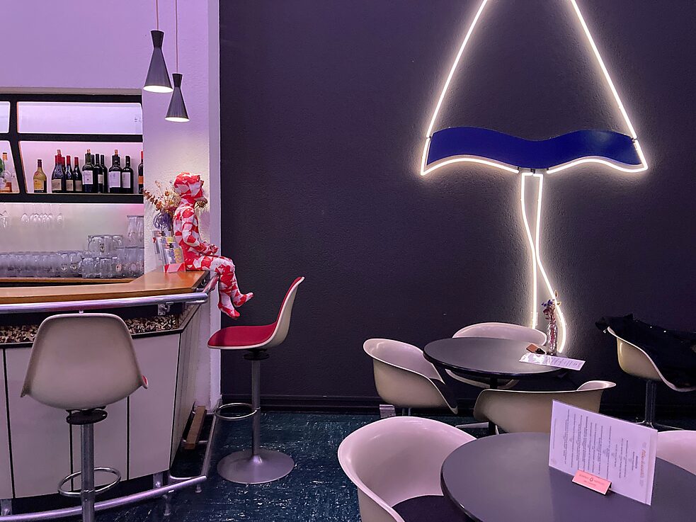 Café: weiße Retrostühle, graue Wand mit einer Leuchtform, schwarze runde Tische, ein blauer Boden