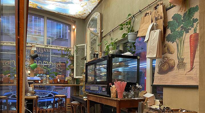 Café corner, on one side a window to the outside, wooden furniture, many plants, a cake counter