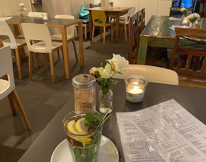 Café: in the foreground a glass with tea and fresh mint and lemon slices, in front of it a small vase with white roses, in the background plain wooden tables and modern white chairs