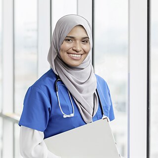 Pflegefachfrau steht in einem modernen Krankenhaus und lacht in die Kamera. Sie trägt Berufskleidung und ein Stethoskop. Sie lacht zufrieden.