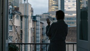 A woman is standing in front of an open window, smoking a cigarette, looking out at illuminated buildings.
