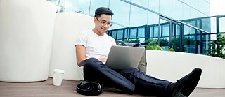 Junger Mann sitzt mit einem Laptop vor einem Firmengebäude