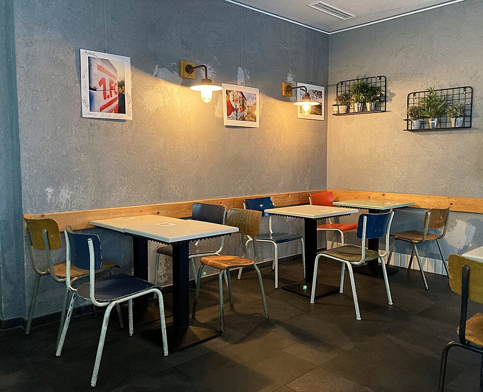 café: graue Wand mit Bildern und Pflanzen, kleine Holztische und bunte Retrometallstühle
