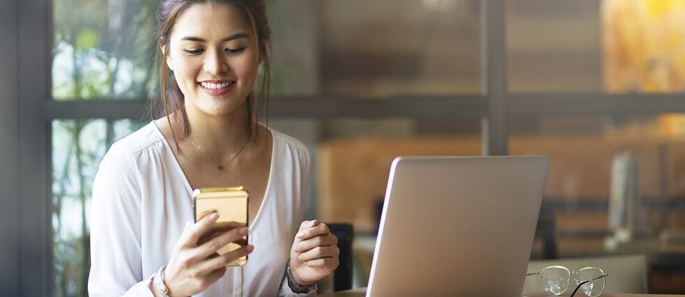 Genç bir kadın ofiste bir masada oturuyor ve bir cep telefonuna bakıyor. Önünde bir laptop bulunuyor.