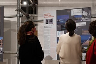 Three visitors are reading and looking at the exhibition board on the Cypriot architects Margarita Danou and Sevina Floridou. The lady on the left wears glasses and a black jumper, the one in the middle wears a loose-fitting white top, and the woman on the right a red jumper with a golden scarf.