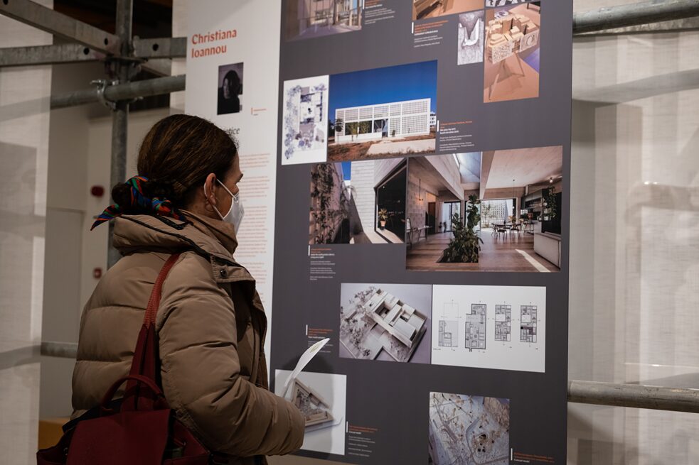 A visitor to the exhibition is looking at a picture of a building designed by Christiana Ioannou. She has an exhibition flyer in her right hand and is wearing a beige jacket, a brown backpack and has a coloured ribbon in her hair.