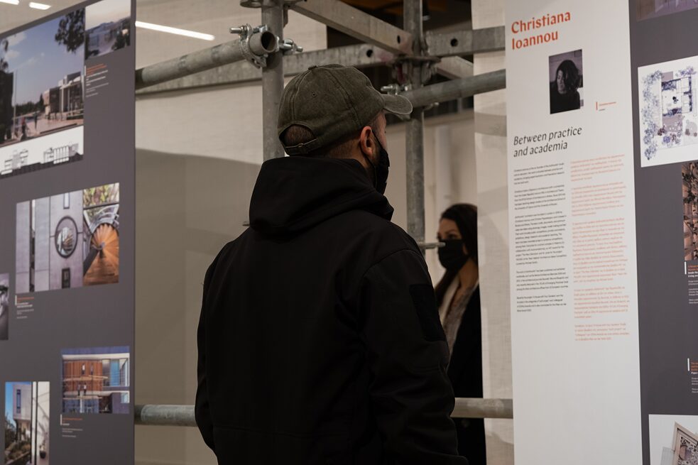 A young man with a face mask, baseball cap and black hoodie reads the text on the exhibition board about the architect Christiana Ioannou. In the background you can see the grey metal scaffolding on which the exhibition panels are mounted and a woman with a black face mask.