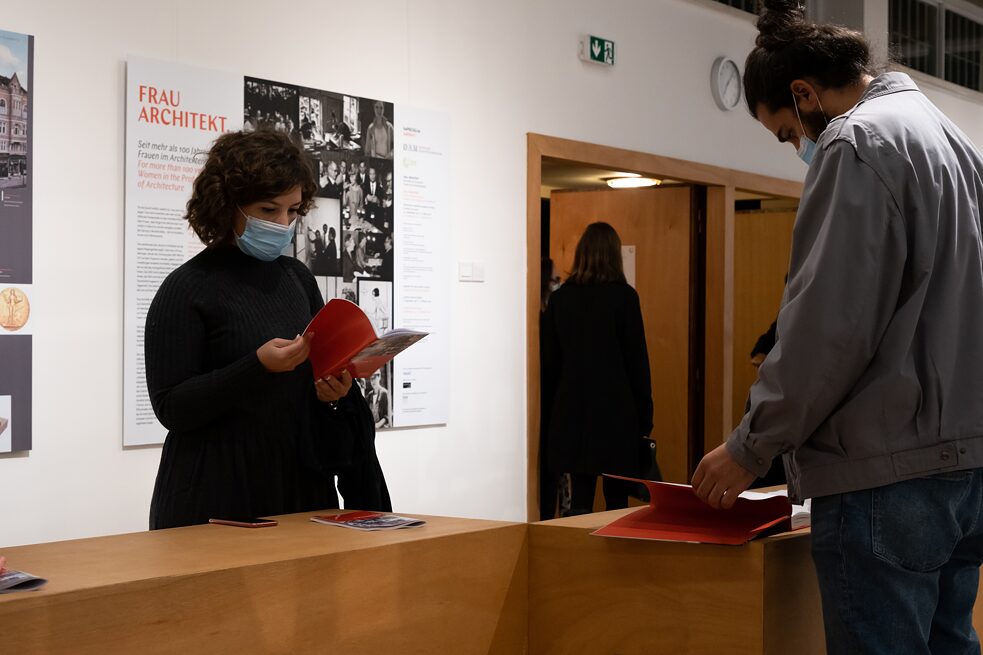 A man in jeans and grey jacket and a women in black clothes wearing face masks are standing in an exhibition room. Each one browses on a booklet. At the background there is a board with the title of the exhibition FRAU ARCHITEKT, text and photos and an open wooden door with people standing around it.