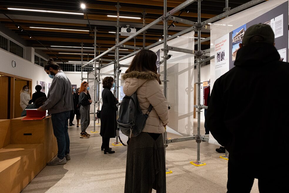 Several visitors wearing face masks are in an exhibition room. In the left half of the picture, a young man in a jeans jacket is browsing through the exhibition catalogue, while in the right half of the picture, several visitors are reading the exhibition panels in concentration.