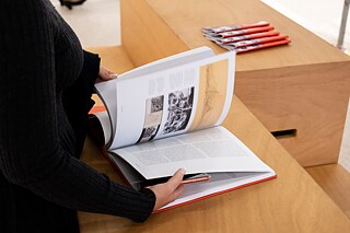 Eine Frau steht an einem Holztisch und durchblättert den Katalog der Ausstellung FRAU ARCHITEKT. Auf dem Tisch liegen weiterhin einige Ausstellungsbüchlein, in welchen Übersetzungen der Ausstellungstexte zu finden sind. Auf der aufgeschlagenen Seite des Kataloges sieht man monochrome Architekturillustrationen.