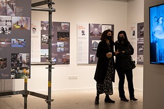 Two ladies are having an intense conversation in the exhibition space. The woman on the left of the picture carries a glass in her right hand and underlines the course of the conversation with a movement of her left hand. In the foreground one can see part of the exhibition scaffolding and a screen. Both visitors are in the back of the photo, dressed in black and wear black face masks.