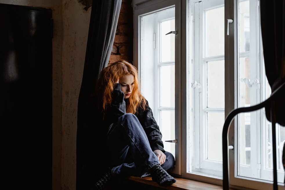 Junge Frau sitzt mit gesenktem Blick am Fenster 